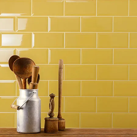 Glanzende gele metrotegels in horizontaal verband als achterwand in een warme keukensetting. Op de voorgrond staan een metalen maatbeker met houten keukengerei en twee houten pepermolens op een rustiek werkblad. De gele tegels zorgen voor een vrolijke, retro en zonnige uitstraling.