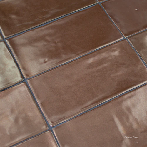 Glanzende koperkleurige wandtegels in langwerpig formaat met onregelmatig, handgemaakt oppervlak. De tegels reflecteren het licht subtiel en tonen lichte kleurnuances voor een warme, luxe uitstraling. Ideaal voor keuken, badkamer of accentmuur in modern of art deco interieur.