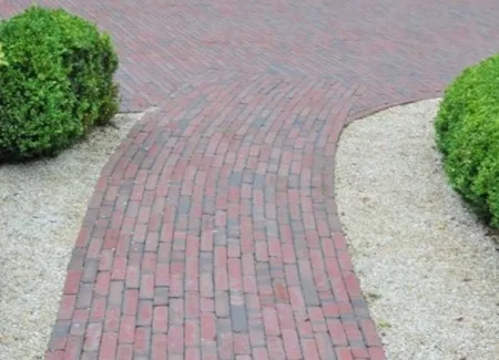 Tuinpad in rode kleiklinkers, aangelegd in een licht gebogen vorm voor een natuurlijke uitstraling. Deze gebakken klinkers zijn kleurvast, slijtvast en bestand tegen intensief gebruik en alle weersomstandigheden. Duurzame inspiratie en advies bij Top Tegel 04 in Ieper, West-Vlaanderen.