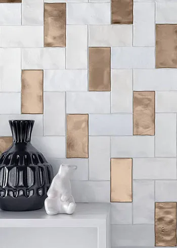 Witte wandtegels in speels visgraatpatroon gecombineerd met glanzende koperkleurige accenten. De mix van matte en blinkende tegels zorgt voor een stijlvol contrast met warme luxe uitstraling. Afgewerkt met zwarte vaas en decoratief beeldje op een minimalistisch wit meubel.